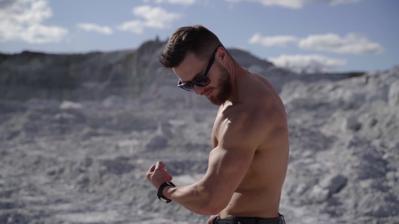 Handsome bodybuilder on the white hill. Strong athlete without shirt shows his arm biceps on nature background. Healthy and sport lifestyle. Slow motion.