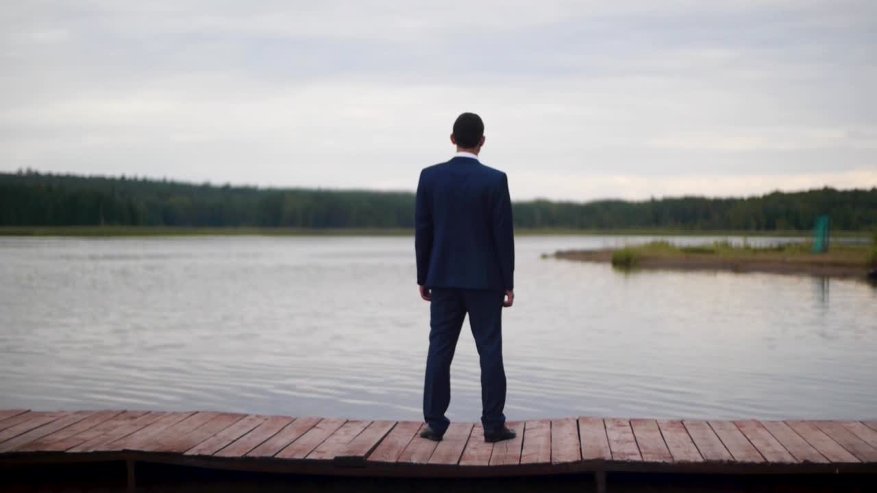 Man in a suit standing on a wooden dock by a lake