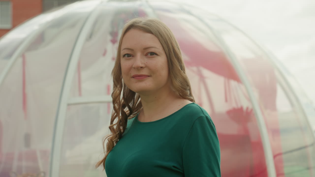 Frau auf einem Dach in der Nähe einer Blasenkapsel, die ein grünes Kleid mit geflochtenem Haar trägt, neben einer Korbschaukel und einer transparenten Kuppel steht, weiches, bedecktes Licht, urbane Skyline im Hintergrund, ruhig und nachdenklich