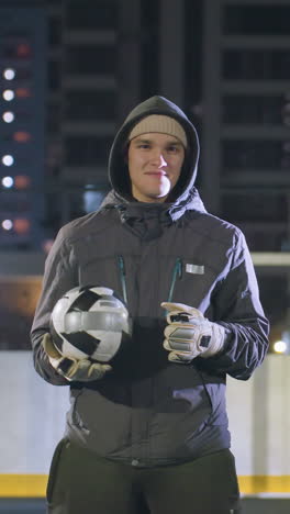 niño joven con capucha con guantes sosteniendo una pelota de fútbol y mostrando una sonrisa sutil en el campo deportivo urbano bajo luces nocturnas, el fondo incluye edificios de gran altura, postes de portería y efectos de luz bokeh
