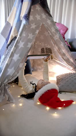 vertical video of african american girl in christmas hat using tablet in blanket tent
