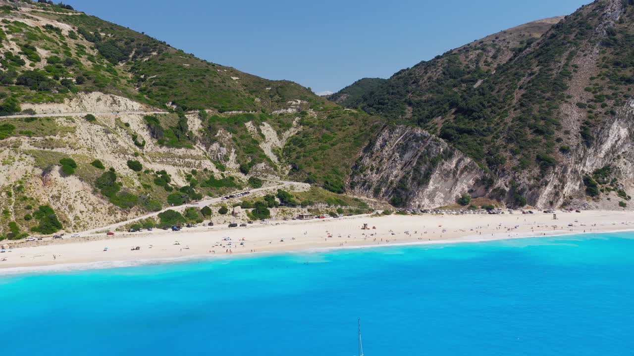 Tourists at Myrtos beach, turquoise water with limestone steep cliffs, hairpin turn road on backdrop, Kefalonia island, Ionian Sea, Aerial revealing shot