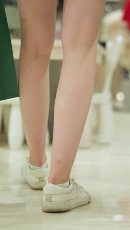 핑크색 쇼트를 입은 여성의 뒷면, 녹색과 빨간색 쇼핑 가방을 들고, 잘 조명된 쇼핑몰의  테이블을 향해 끔하게 배열된 옷을 걸어다니고, 다른 쇼핑객들과 함께