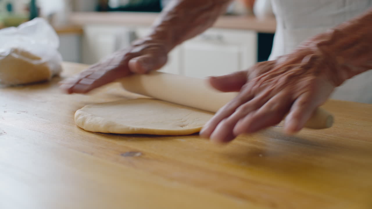 Hands of Senior Woman Flattening Dough with Rolling Pin on Wooden Table