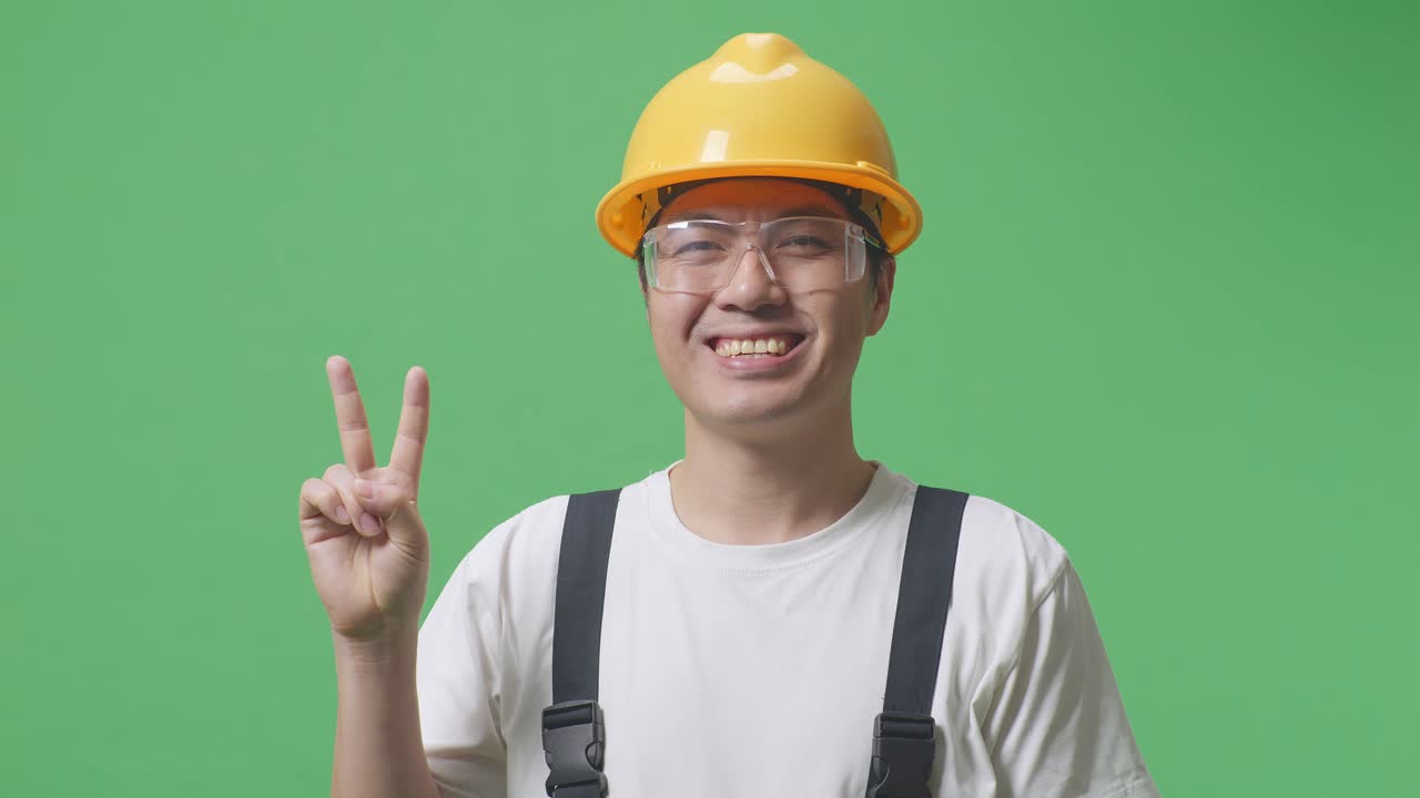 Close Up Of Asian Man Worker Wearing Goggles And Safety Helmet Smiling And Showing Peace Gesture To Camera While Standing In The Green Screen Background Studio