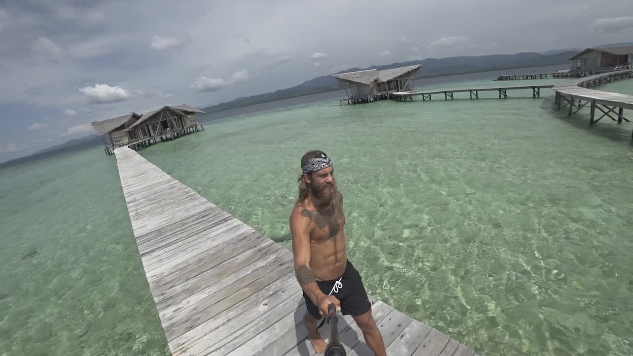un hombre joven, en forma y fuerte con pelo largo y barba camina por el embarcadero y gira para mostrar el hermoso entorno del complejo de lujo con bungalows individuales en el agua