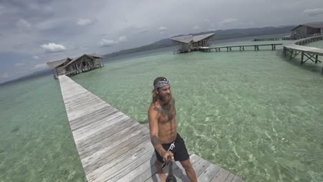 un hombre joven, en forma y fuerte con pelo largo y barba camina por el embarcadero y gira para mostrar el hermoso entorno del complejo de lujo con bungalows individuales en el agua