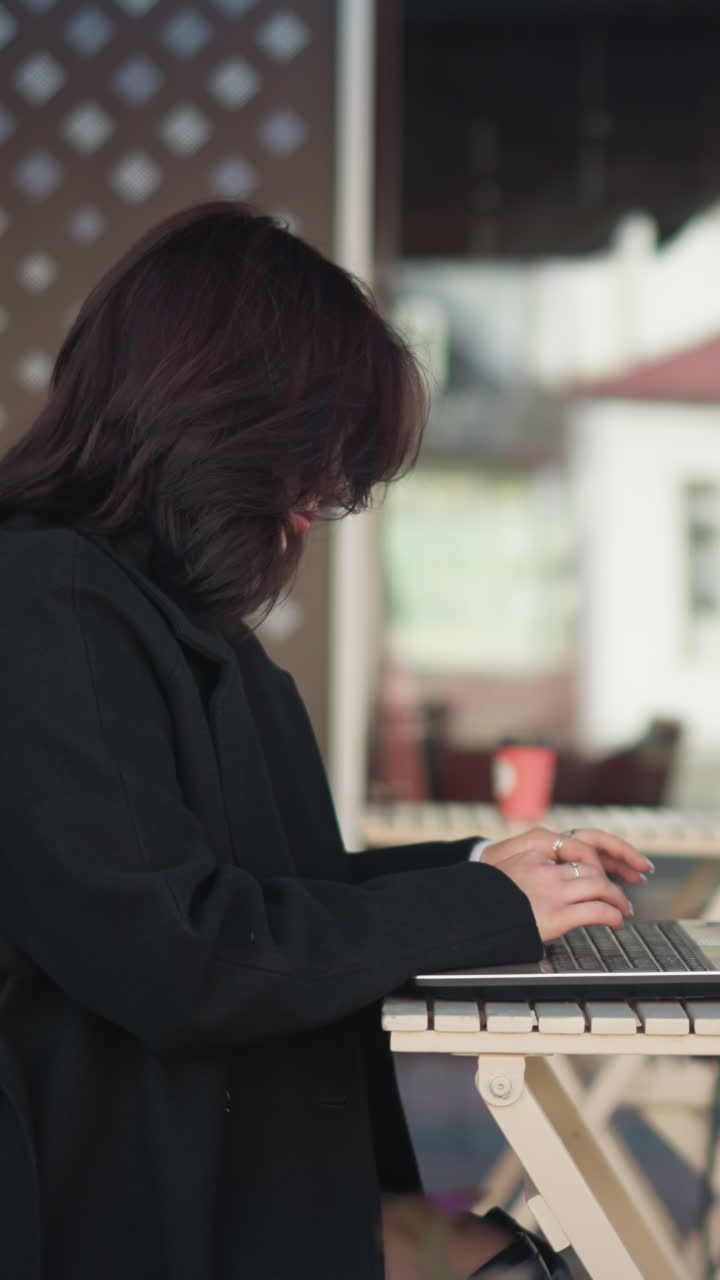 도시 환경에서 현대 가구 근처에 앉아, 뒷면에 반사되는 유리 건물, 꽃과 함께 야외에서 노트북에서 일하는 여성의 측면 뷰