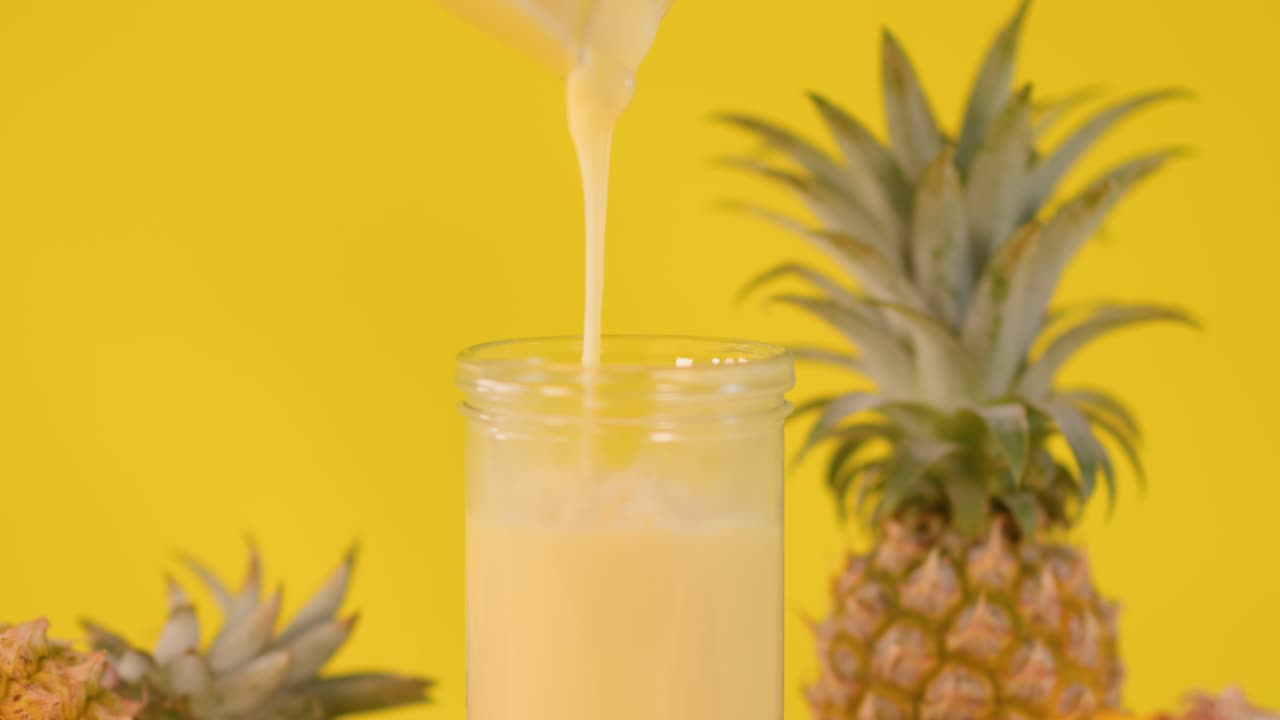 Creamy pineapple smoothie poured into glass, surrounded by fresh pineapples, bright yellow background, static shot