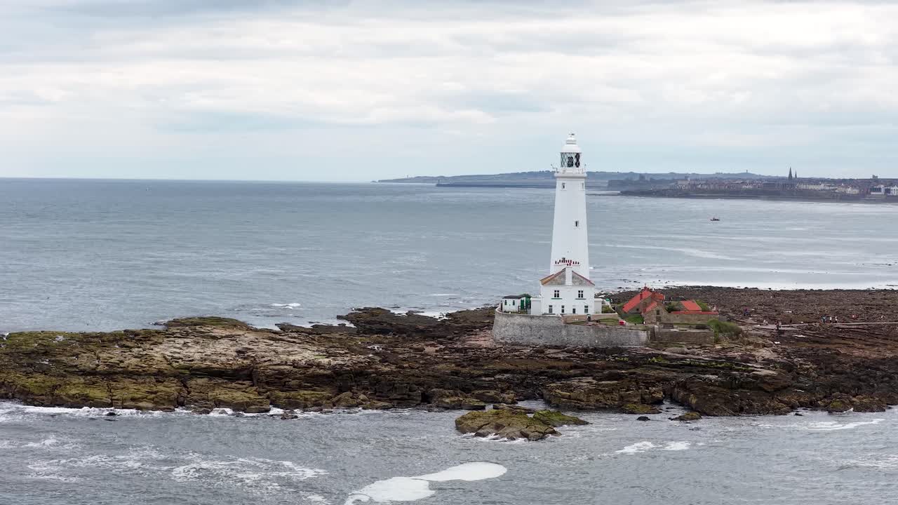 Aerial footage pans across a white lighthouse on a rocky shoreline, with overcast skies and calm sea, highlighting coastal scenery and maritime architecture