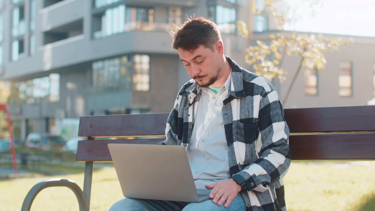 Mature businessman on video call using laptop while sitting on bench in downtown city street