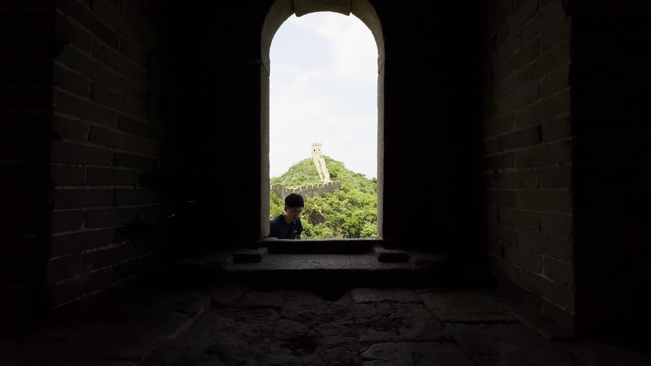 View from inside a watchtower on the Great Wall of China as a man enters through a small window, showcasing the Great Wall behind