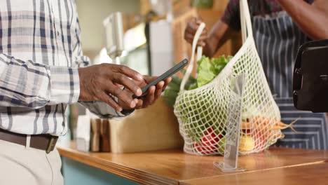 Midsection-of-senior-african-american-man-paying-using-smartphone-at-health-food-shop,-slow-motion