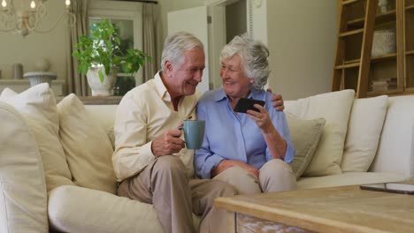 Kaukasisches-älteres-Paar-Lächelt,-Während-Es-Gemeinsam-Sein-Smartphone-Benutzt-Und-Zu-Hause-Auf-Der-Couch-Sitzt