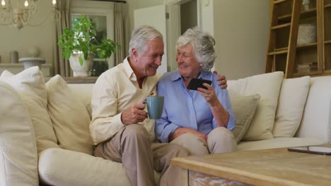 caucasian senior couple smiling while using smartphone together sitting on the couch at home