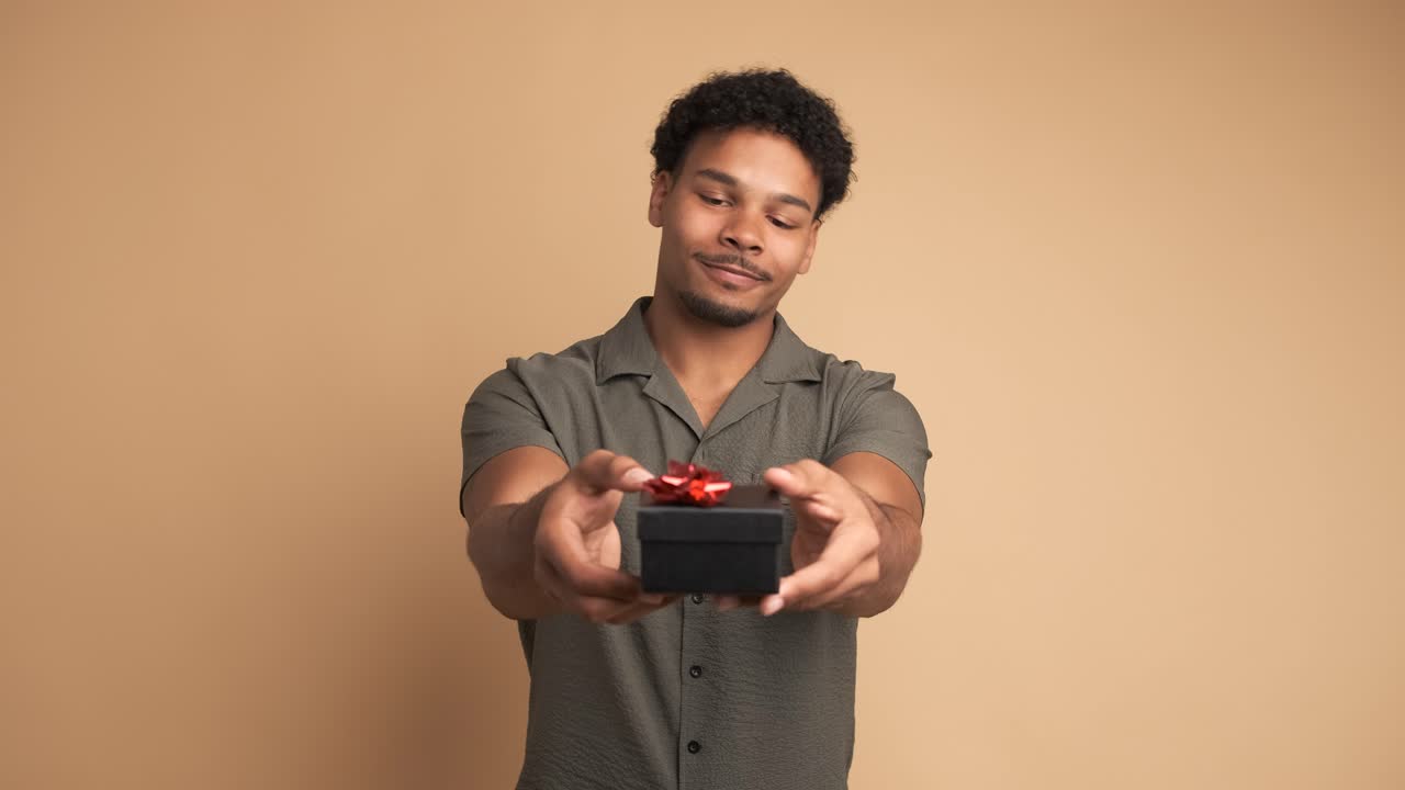 Happy man giving gift box and looking at camera in beige studio
