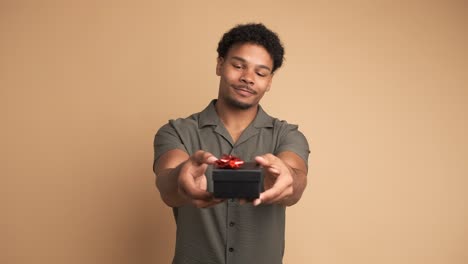 happy man giving gift box and looking at camera in beige studio
