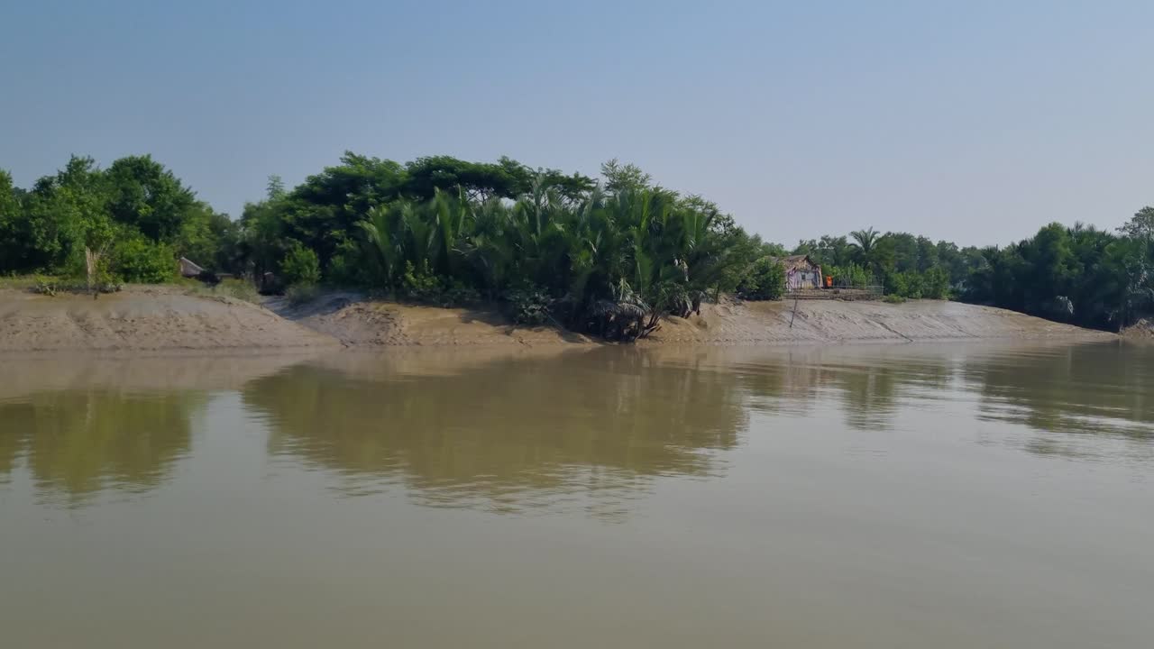 Vista dal punto di osservazione di un passeggero a bordo di una barca che naviga lentamente su un fiume delle Sundarbans in Bangladesh, sotto un cielo soleggiato, con la bassa marea che rivela la costa fangosa e la vegetazione tropicale, patrimonio dell'UNESCO