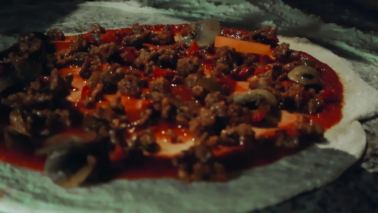 Close up view of a chef or a home cooks diverse hand topping a home made pizza dough with tomato sauce and minced meat toppings in slow motion. The dough is freshly made, white flour on the table