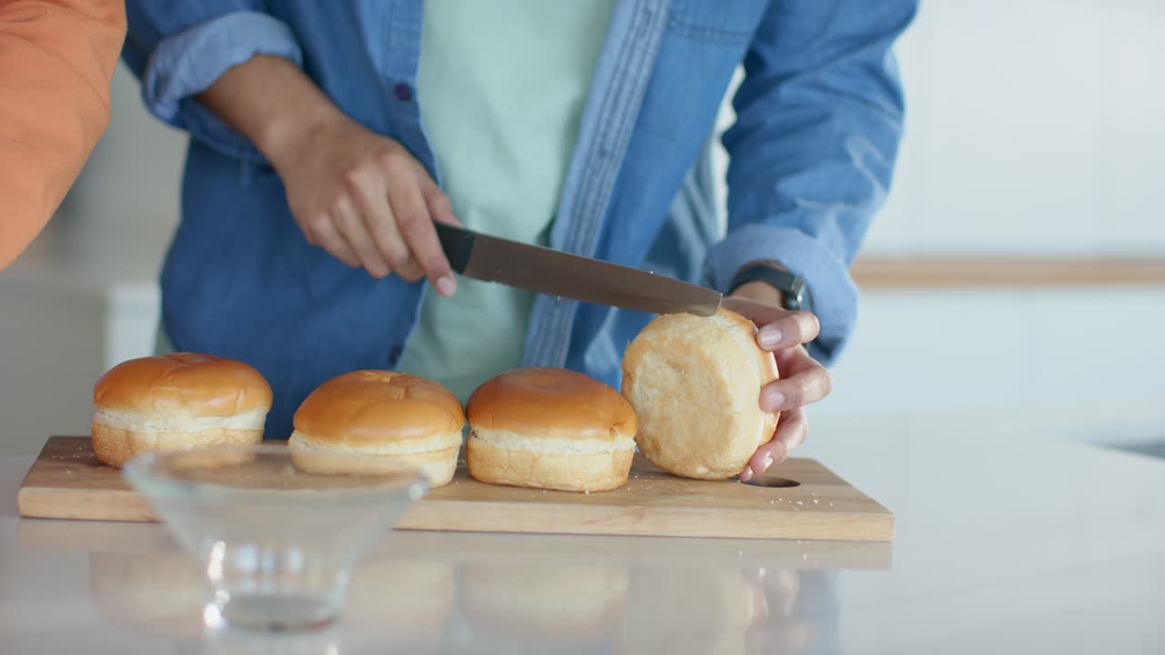 Mand i denim deler bolle med kniv ved køkkenbordet og forbereder sandwich efter at have skåret tomater.