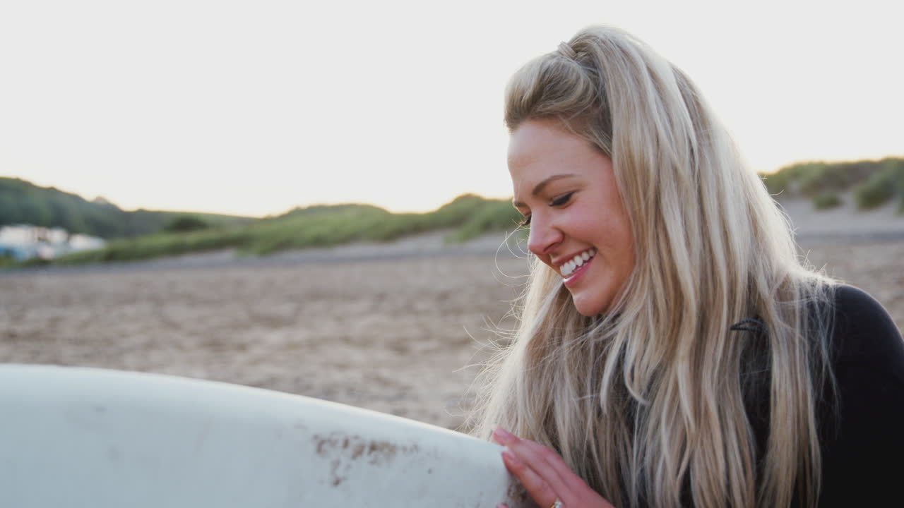 해가 지는 동안 해변에서 서핑 휴가를 즐기는 서핑 보드를 들고 있는 웨이트 슈트를 입은 여성