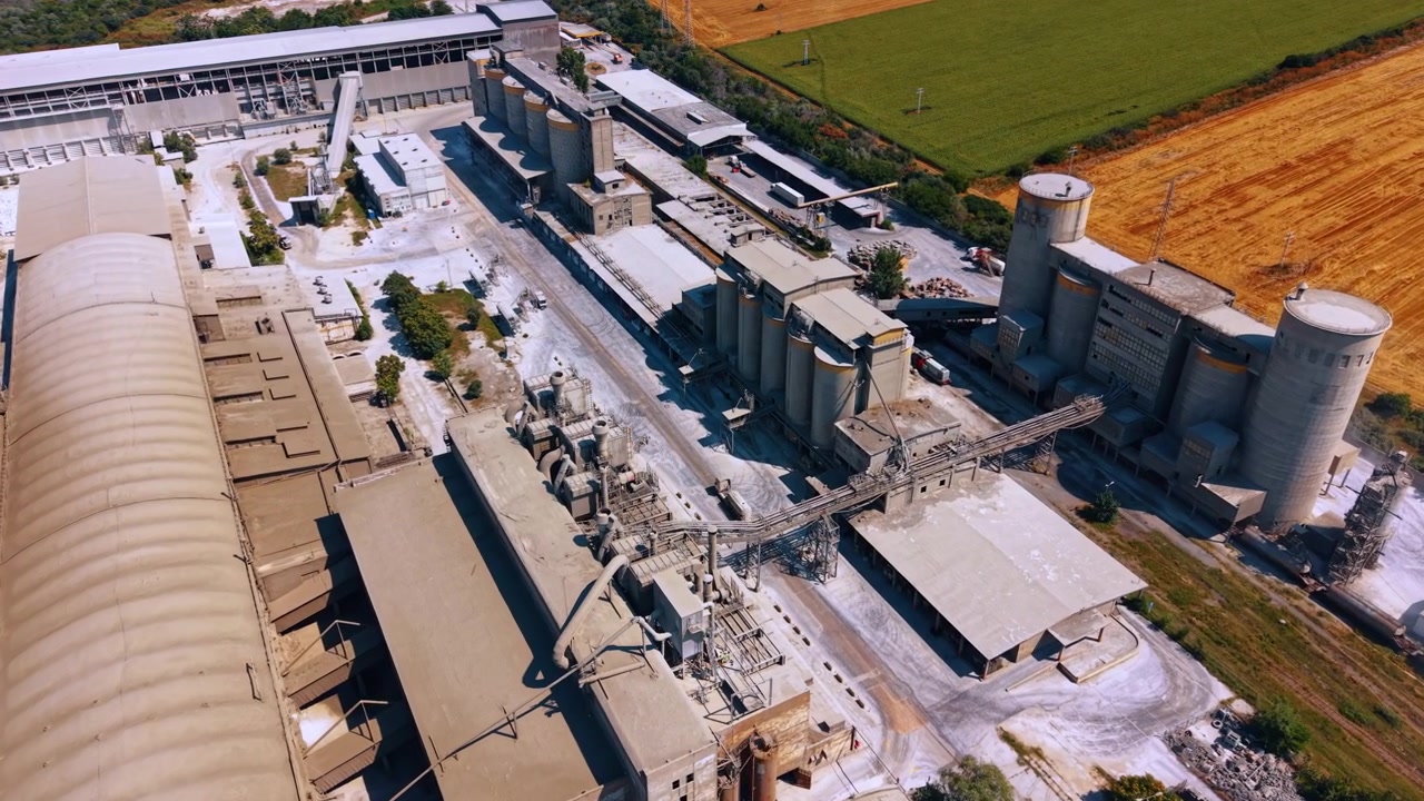 Large industrial cement plant with multiple storage silos. The high angle drone perspective shows the factory complex adjacent to agricultural fields.