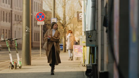 Hübsche-Elegante-Frau-In-Beigem-Mantel,-Die-Alleine-Die-Städtische-Geschäftsstraße-Entlang-Läuft,-Elektroroller-Auf-Dem-Bürgersteig