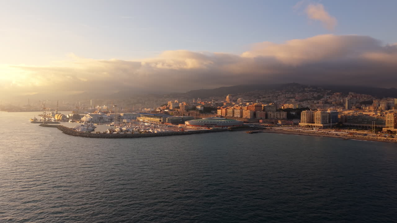 Drone che vola sul mare verso il porto di Genova con edifici cittadini e calda luce del tramonto sulla costa