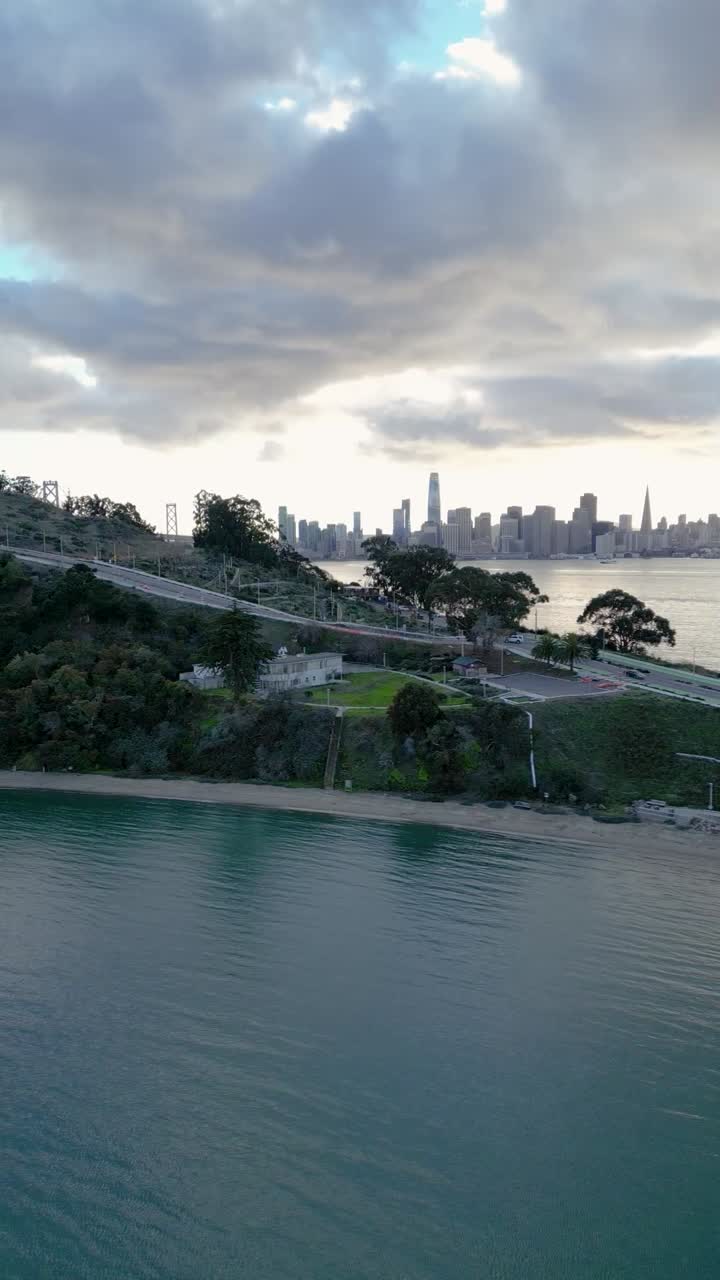 Side-tracking drone shot of Yerba Buena Island with San Francisco's skyline visible across the bay, highlighting both natural and urban landscapes.