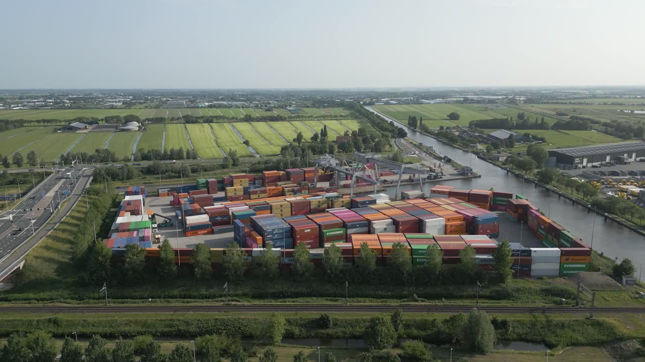 High-resolution drone footage showing a large container terminal filled with vibrant shipping containers beside a canal, surrounded by typical Dutch farmland