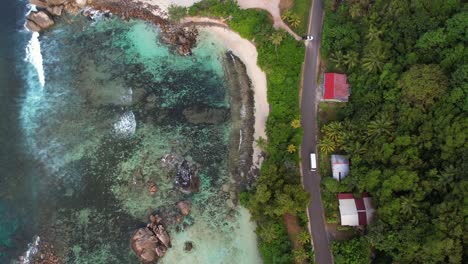 Bird-eye-drone-shot-of-road,-passing-vehicle-near-the-beach-of-Port-glaud-Mahe-Seychelles