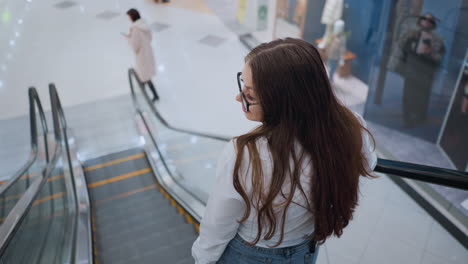 back view of young adult with long, flowing hair descending escalator in busy mall, she elegantly rests her hand on rail while observing surroundings, with people and storefronts visible