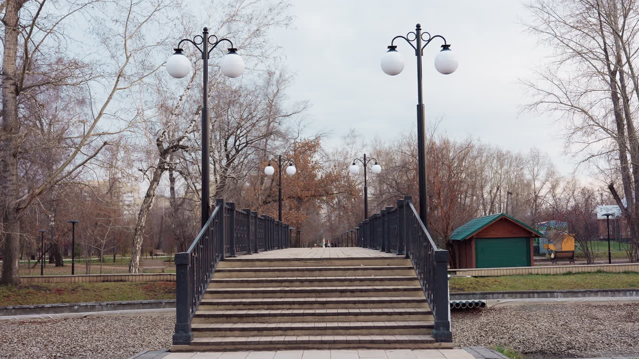 Pedestrian bridge with stairs in winter park surrounded by leafless trees and lampposts, small green-roofed wooden house beside pathway adds rustic touch to calm outdoor urban setting