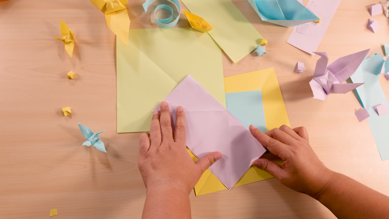 Person folds pastel origami paper, surrounded by finished models, under soft, even lighting