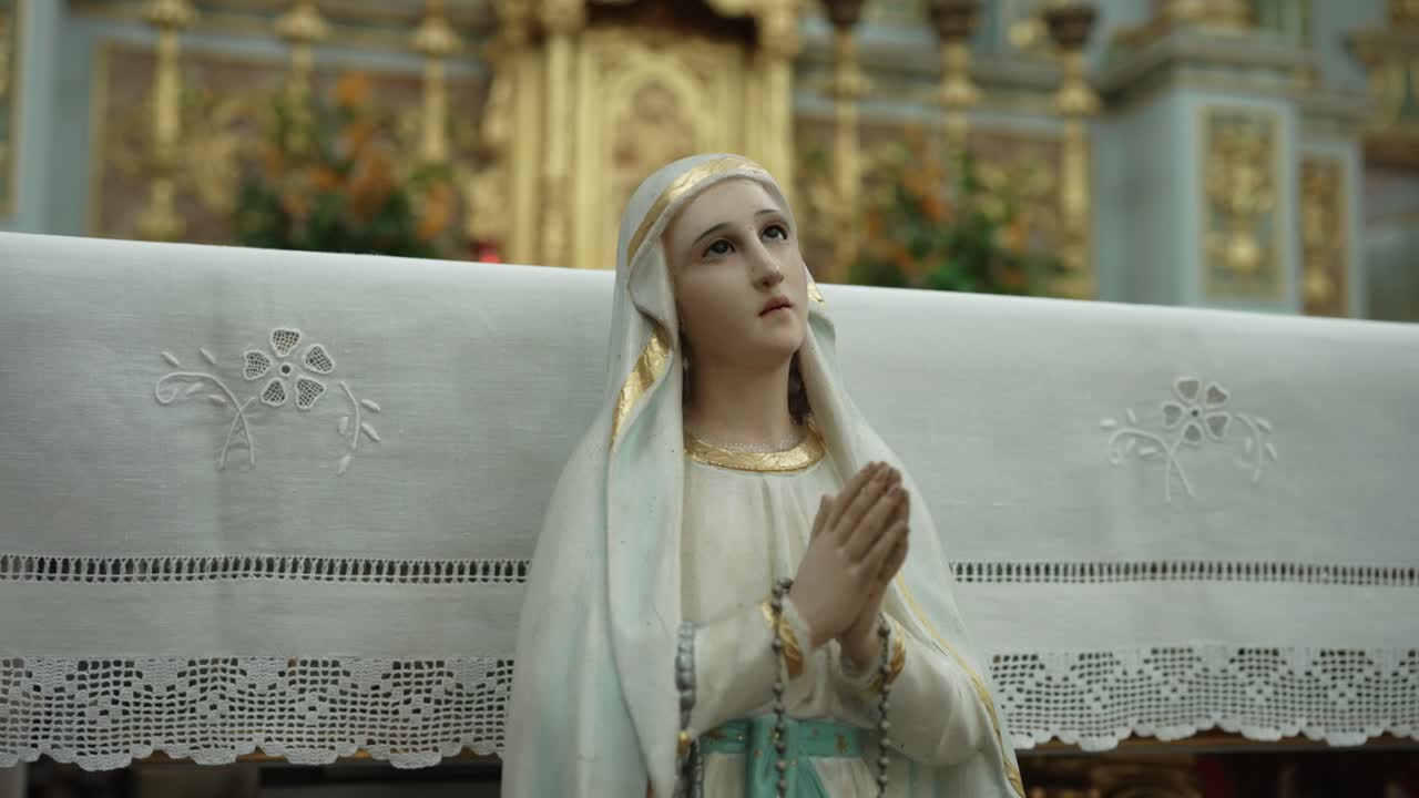 close up of Virgin Mary statue with folded hands and rosary before ornate altar
