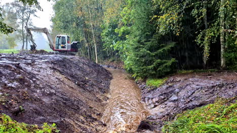 an excavator sits above the muddy stream as water flows past a wooden barrier and green vegetation - pan down shot