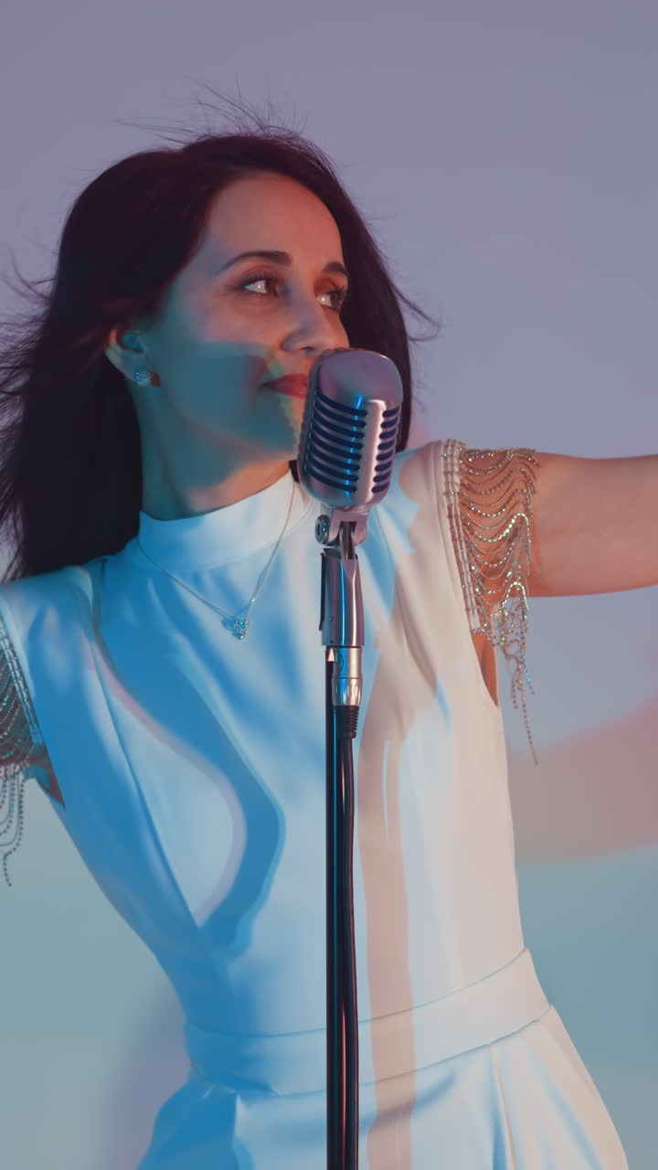 Singer in a white gown with eyes closed, moving hands gracefully in the air while performing into a vintage microphone. Set against a soft gradient background of blue and pink