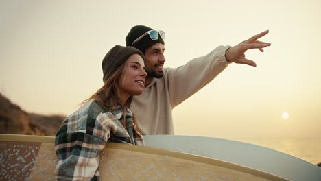 A-brunette-man-in-a-black-hat-and-white-top-points-somewhere-in-the-direction-of-his-blonde-girlfriend-in-a-plaid-shirt-while-they-stand-on-the-seashore-at-dawn-in-the-morning-and-hold-surfboards-in-their-hands