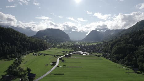 Frontales-Drohnenvideo,-Das-Sich-über-Farmen-Und-Häuser-Im-Triglav-Bewegt,-Mit-Hügeln-Am-Horizont,-Der-Sonne-Im-Vordergrund-Und-Einem-Klaren-Himmel-Mit-Wolken