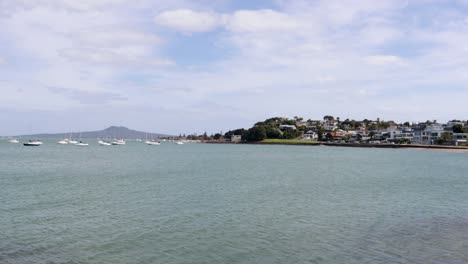 A-handheld-shot-of-a-lake-with-blue-water-and-some-boats-floating-on-it,-with-a-neighborhood-and-a-volcano-in-the-background-on-a-cloudy-morning-in-the-city-of-Auckland,-New-Zealand