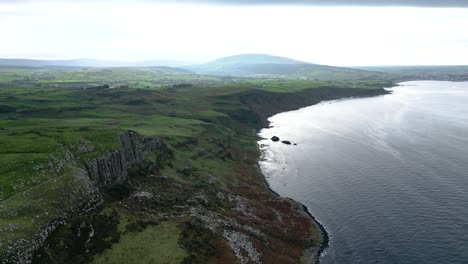 Vista-Aérea-Amplia-De-La-Cabeza-De-Feria-Geográficamente-Prominente-En-Irlanda-Del-Norte,-Conocida-Por-Sus-Impresionantes-Acantilados-Costeros-Con-Vistas-Al-Mar-Tranquilo-Que-Refleja-Y-Al-Gran-Paisaje-Con-Montañas