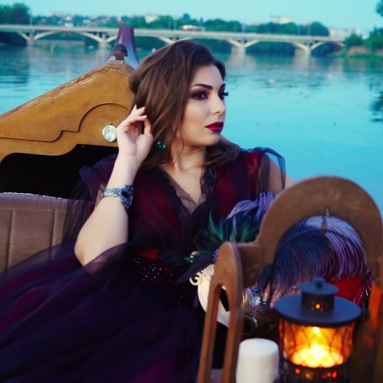 A woman with make-up is sitting in the gondola and holding mask in her hands with a colored feathers near a decorative lantern and white candles on the background of the river in the evening. Carnival in Venice