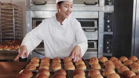 portrait of happy asian female baker working in bakery kitchen, placing rolls on trays, slow motion