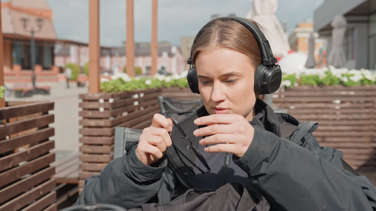 Giovane donna caucasica seduta su una panchina che apre il suo zaino mentre indossa delle cuffie, tira fuori degli occhiali da sole accanto a una tazza di caffè da asporto, terrazza soleggiata con listelli di legno e skyline della città in lontananza, concentrata e curiosa
