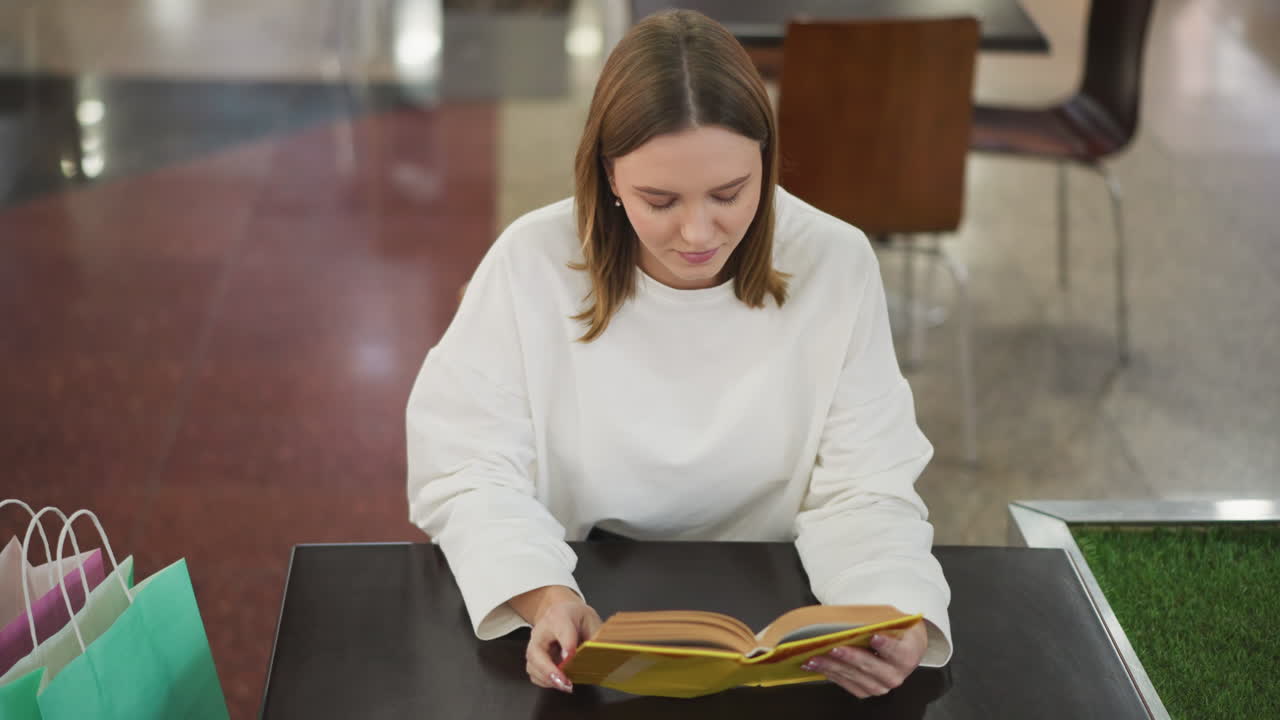 joven leyendo un libro amarillo en una mesa en un restaurante moderno con bolsas de compras a su lado, parece centrada y contenta en el vibrante entorno interior