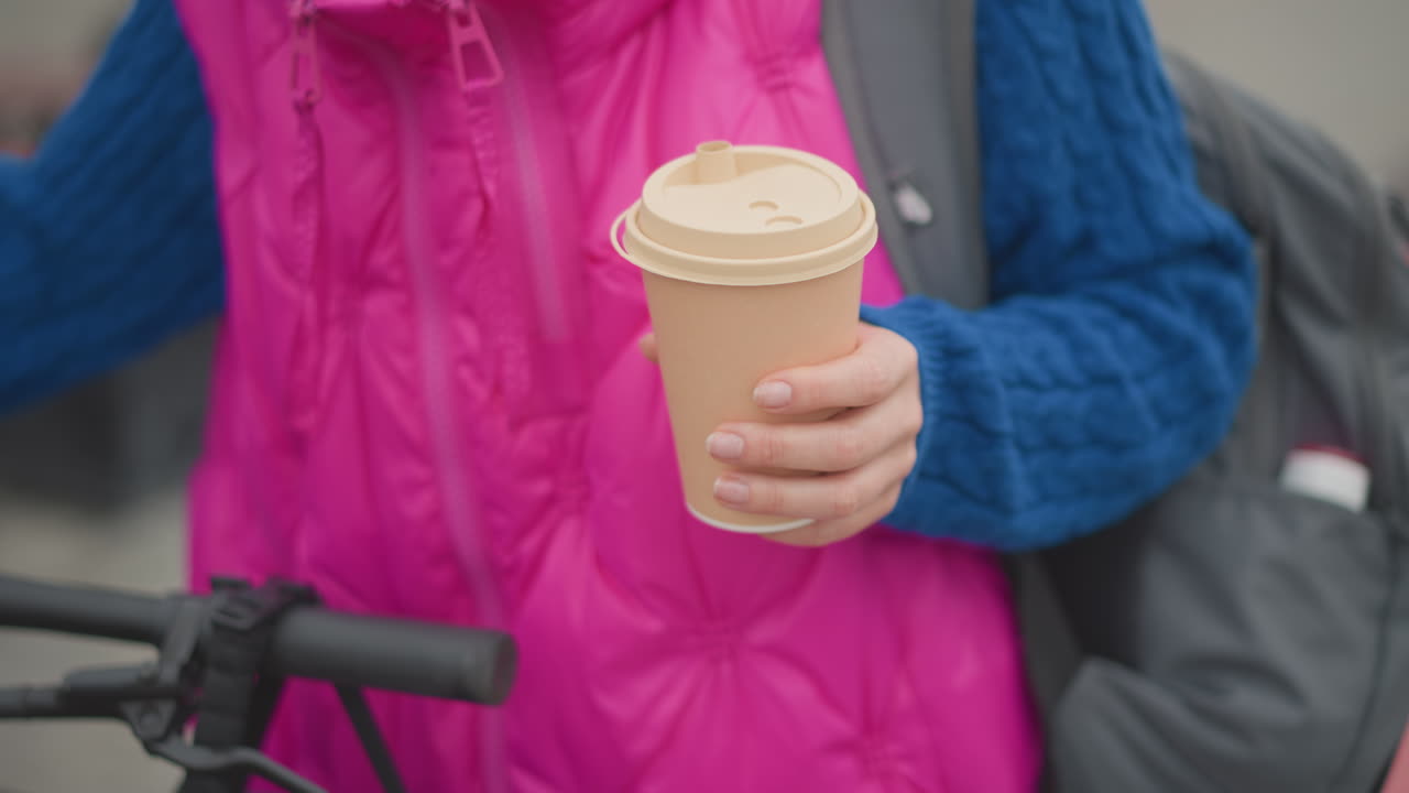 Frau mit Kaffee in der Hand in der Nähe eines Fahrradlenkers, pinke Weste und blauer Pullover, Rucksack auf der Schulter, Nahaufnahme eines Coffee-to-go-Bechers, morgendlicher Arbeitsweg in der Stadt, sanftes Sonnenlicht, verschwommener Stadthintergrund, gemütliche, einsame Stimmung