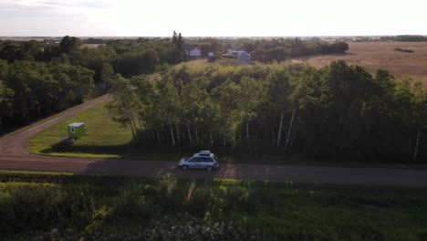 toma aérea de 4k de una minivan plateada conduciendo lentamente por un camino de tierra junto a los árboles durante la puesta de sol en un entorno rural