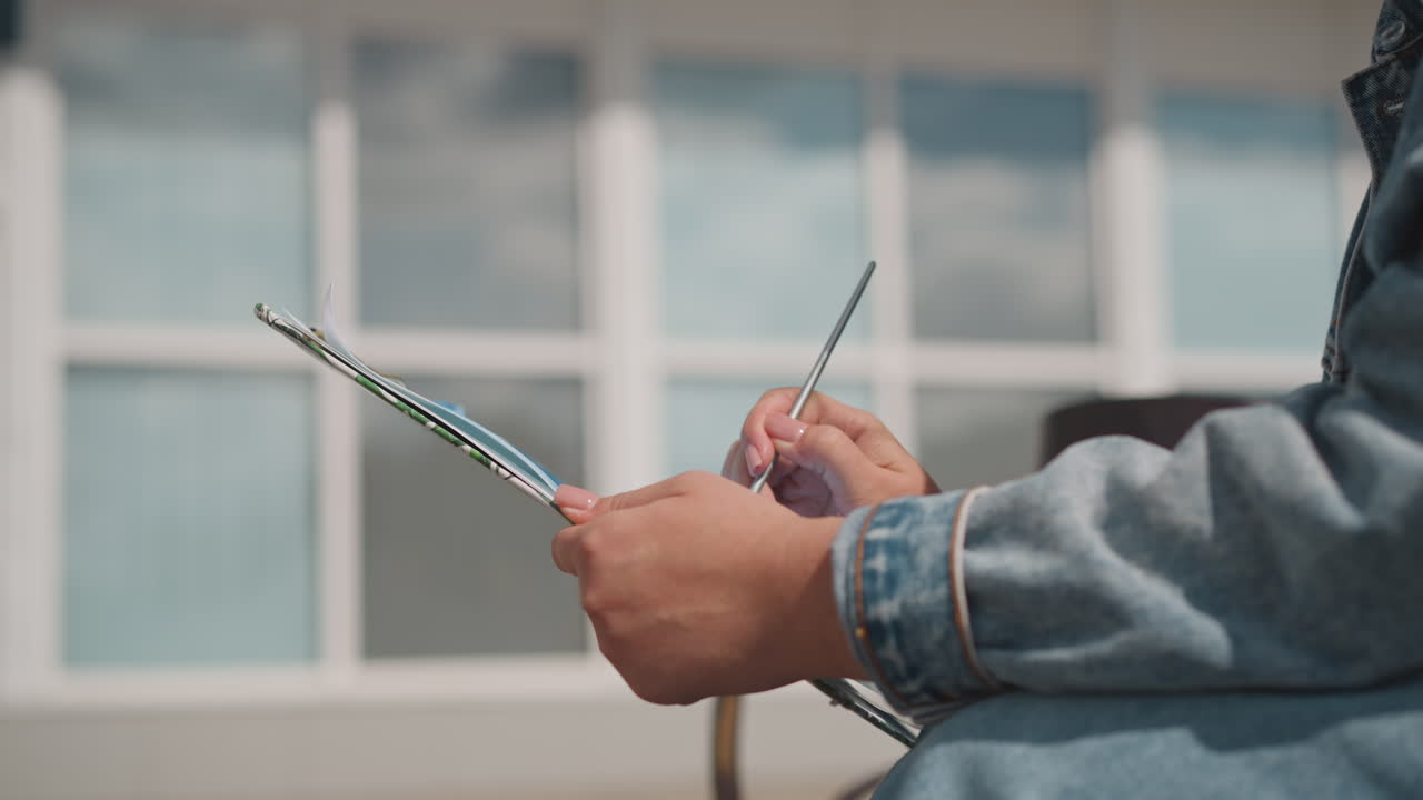 Handheld palette and brush by window, denim jacketed artist mixes paint while seated on bench, urban facade background with soft grid windows, sunlight highlights brush movement, candid editorial mood