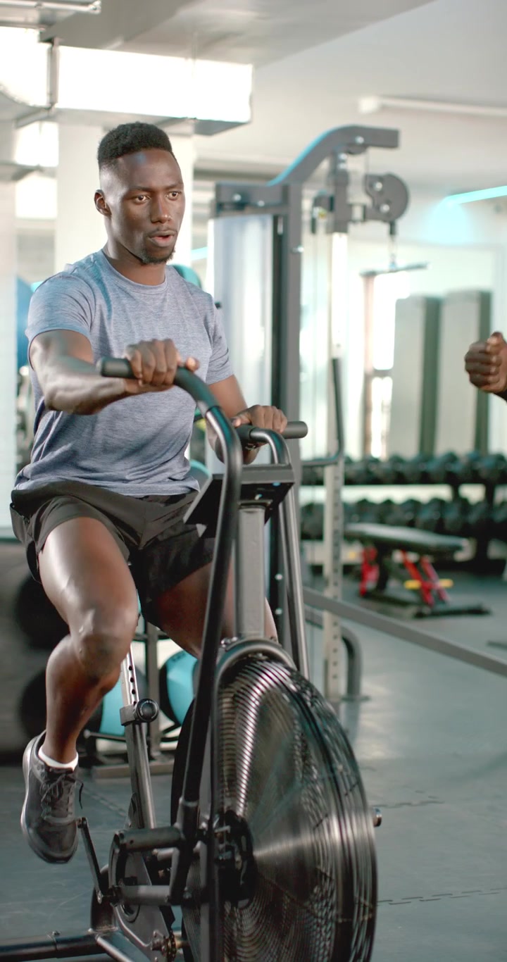 Vertical video: Trainer timing African American man pedaling air bike for interval training at gym