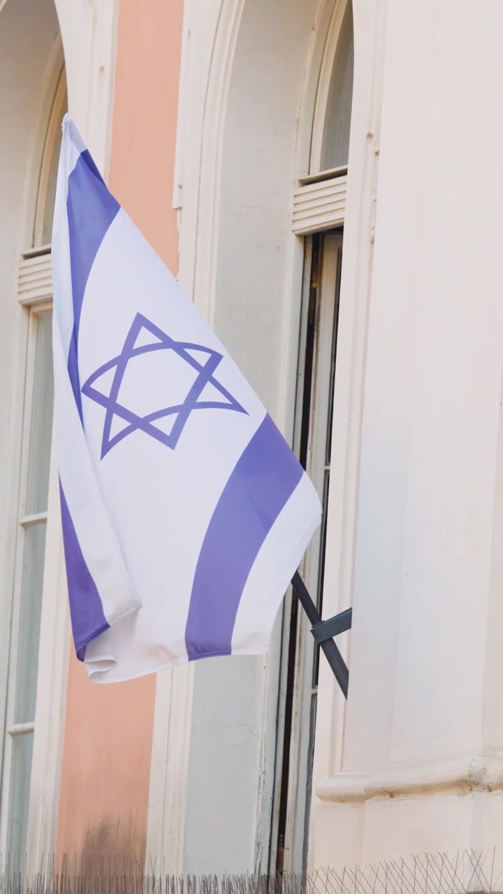 Bandiera israeliana sventola su un'asta, luce diurna intensa, finestra ad arco storica, facciata di un edificio dai colori pastello tenui